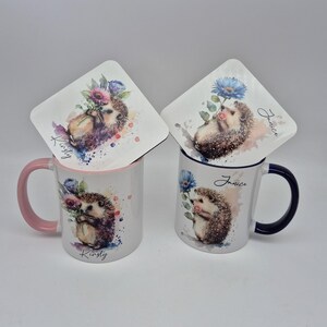 May include: Two ceramic mugs and coasters featuring watercolor hedgehog designs. One mug has a pink handle and rim, the other has a dark blue handle and rim. The coasters and mugs have the names "Kirsty" and "Janice" printed on them.