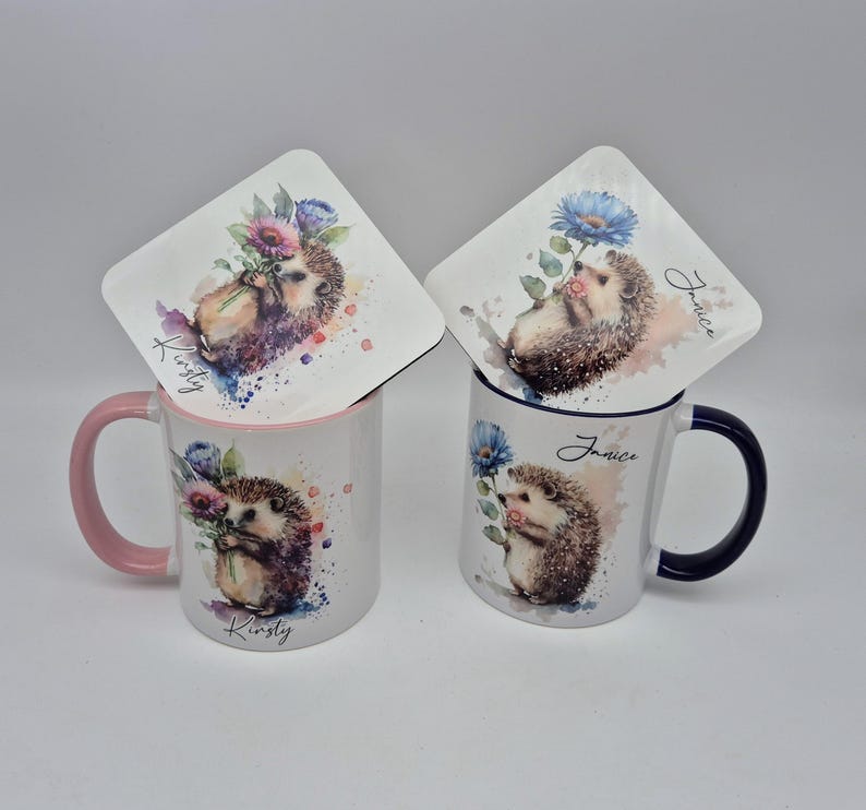 May include: Two ceramic mugs and coasters featuring watercolor hedgehog designs. One mug has a pink handle and rim, the other has a dark blue handle and rim. The coasters and mugs have the names "Kirsty" and "Janice" printed on them.