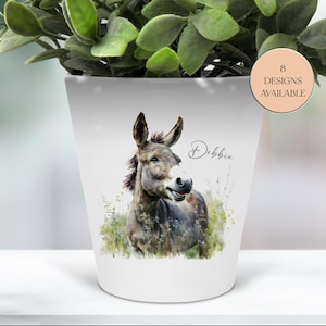 以下が含まれることがあります： 白いセラミック植木鉢。水彩画で描かれたロバのイラストが特徴です。ロバはグレーと茶色の色合いで描かれ、笑顔を浮かべています。「Debbie」という名前が筆記体で書かれています。円形のラベルには「8 DESIGNS AVAILABLE」と記載されています。