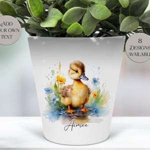 May include: White ceramic plant pot with a watercolour illustration of a yellow duckling in water, surrounded by blue and yellow flowers. The pot is inscribed with the name "Aimee". Text on the pot says "Add your own text" and "8 designs available".