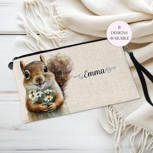 May include: A beige zippered pouch featuring a watercolor illustration of a squirrel holding daisies. The name "Emma" is written in script. A small pink circle indicates "8 DESIGNS AVAILABLE". The pouch has a black wrist strap.