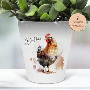May include: White ceramic planter featuring a watercolor illustration of a rooster in shades of brown, red, and blue. The name "Debbie" is written in cursive. A circular label indicates "7 DESIGNS AVAILABLE". The planter is filled with green foliage.