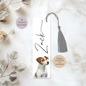 May include: A white bookmark with a Jack Russell Terrier illustration and the name "Zack" in script. A grey tassel is attached. The text "8 designs available" and "Tassel colours at random" are also visible. A decorative item for book lovers.