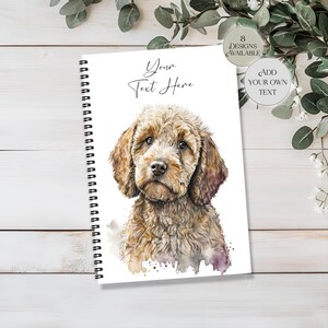 May include: A spiral-bound notebook with a watercolour illustration of a brown and tan Goldendoodle dog. The cover has the text "Your Text Here" in script. Additional text indicates "8 Designs Available" and "Add Your Own Text".