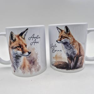 May include: Two white ceramic mugs with a watercolor fox design. One mug has the text "Auntie Ann" and the other has "Uncle Brian". The foxes are depicted in shades of brown, orange, and white.