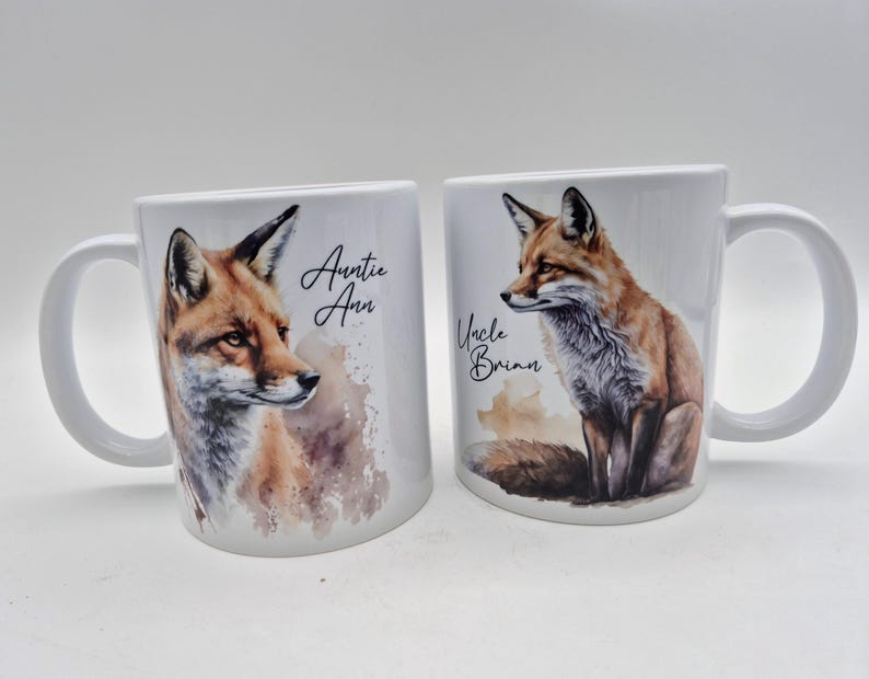 May include: Two white ceramic mugs with a watercolor fox design. One mug has the text "Auntie Ann" and the other has "Uncle Brian". The foxes are depicted in shades of brown, orange, and white.