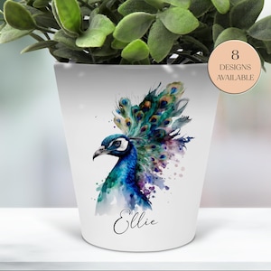 May include: White ceramic planter with a watercolor peacock design in shades of blue, teal, and purple. The planter has the name "Ellie" in script and a circular label that says "8 designs available". The planter is filled with green leafy plants.
