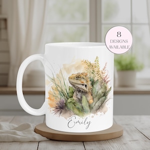 May include: White ceramic mug with a watercolour illustration of a bearded dragon amongst foliage. The mug has a curved handle and the name "Emily" is written in cursive. A pink circle indicates "8 designs available".