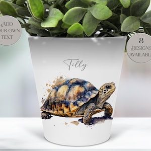 May include: White ceramic plant pot with a tortoise watercolour illustration. The pot is inscribed with the name "Tilly" and the text "Add Your Own Text" and "8 Designs Available" on circular badges. A green plant sits inside the pot.