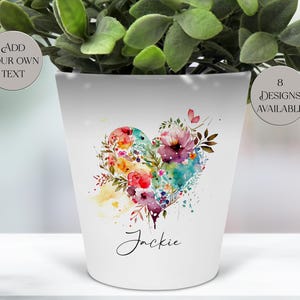 Peut inclure: Pot de fleurs en céramique blanche avec un motif de cœur à l'aquarelle rempli de fleurs colorées. Le pot porte le nom "Jackie" écrit en cursive. Le texte sur le pot indique "Ajoutez votre propre texte" et "8 modèles disponibles".