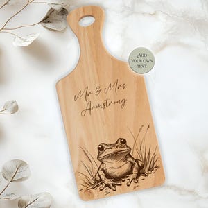 Könnte beinhalten: Ein hölzernes Schneidebrett mit Griff, graviert mit den Worten "Mr & Mrs Armstrong" und einer detaillierten Illustration eines Frosches. Das Brett ist hellbraun und hat eine natürliche Holzmaserung.
