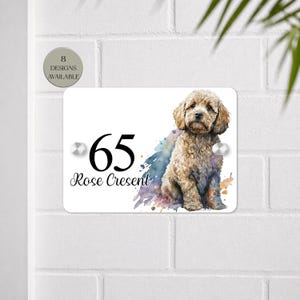 Peut inclure: Panneau de numéro de maison rectangulaire blanc avec une illustration de chien à l'aquarelle. Le panneau affiche le numéro "65" en noir et "Rose Crescent". Le panneau est monté sur un mur de briques blanches.