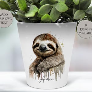 May include: White ceramic plant pot with a watercolour illustration of a sloth. The sloth has brown and tan fur. The pot has the text "Aimee" in script and the text "Add Your Own Text" and "8 Designs Available" in circles.