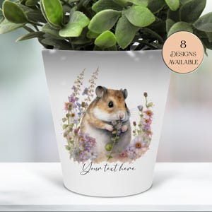 May include: White ceramic planter featuring a watercolor illustration of a hamster surrounded by purple and pink flowers. The planter has the text "Your text here" at the bottom and a circular graphic that says "8 Designs Available".