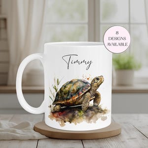 Könnte beinhalten: Weiße Keramiktasse mit einer Aquarellillustration einer Schildkröte mit buntem Panzer. Die Tasse trägt den Namen "Timmy" in Schreibschrift und eine Grafik mit der Aufschrift "8 Designs Available". Die Tasse steht auf einem Holzuntersetzer.