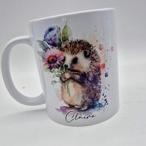 May include: White ceramic mug with a watercolour illustration of a hedgehog holding flowers. The design includes purple, pink, and blue floral elements, with the name "Claire" in script. The mug has a curved handle.
