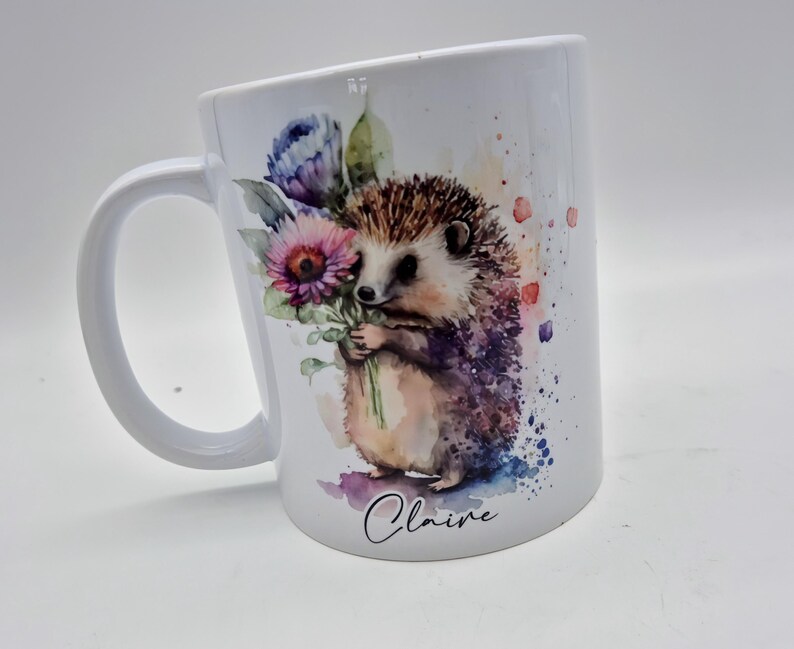 May include: White ceramic mug with a watercolour illustration of a hedgehog holding flowers. The design includes purple, pink, and blue floral elements, with the name "Claire" in script. The mug has a curved handle.