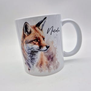 May include: White ceramic mug featuring a watercolor illustration of a fox with orange, brown, and gray fur. The mug has a curved handle and the name "Nicola" is written in black script.