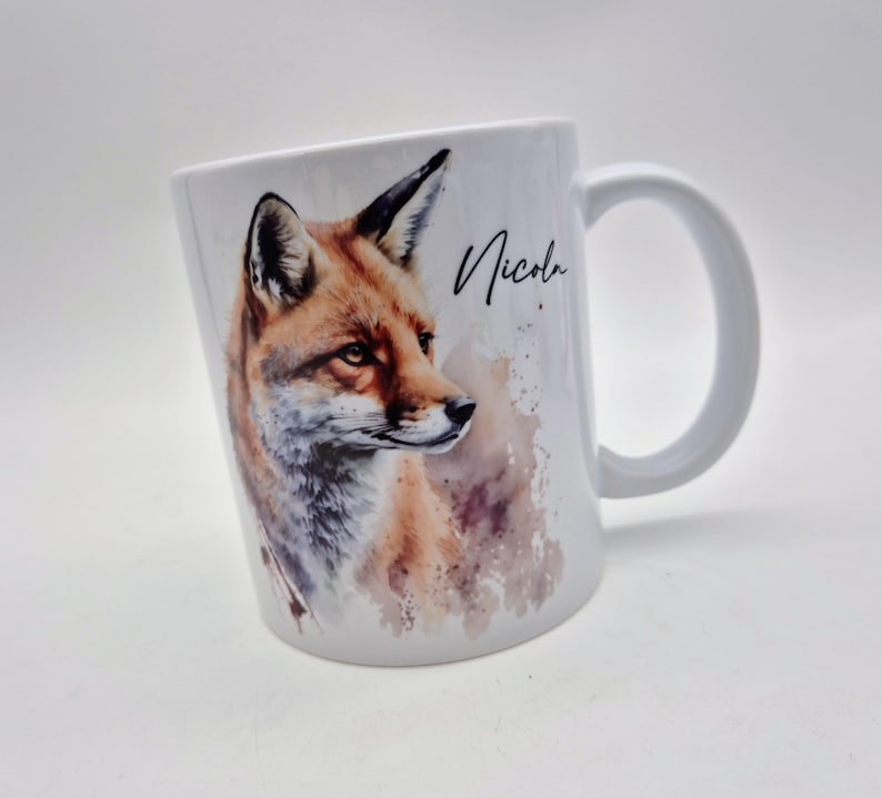 May include: White ceramic mug featuring a watercolor illustration of a fox with orange, brown, and gray fur. The mug has a curved handle and the name "Nicola" is written in black script.