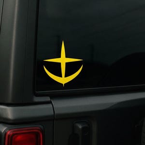 以下が含まれることがあります： 黒いSUVの後部窓に貼られた、黄色いアンカーデザインのステッカー。上部に十字架が付いています。ステッカーはシンプルなグラフィックデザインです。車両のテールライトが見えます。