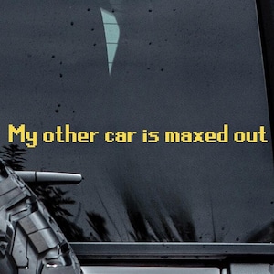 May include: A close-up of the rear of a silver SUV with a black spare tire and a black window. The text "My other car is maxed out" is in yellow on the window.