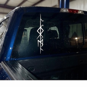 May include: White vinyl decal featuring a vertical runic symbol, affixed to the rear window of a blue pickup truck. The symbol is composed of angular lines and geometric shapes, creating a striking contrast against the dark background.