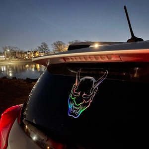 Może przedstawiać: Srebrny samochód z tęczową, opalizującą naklejką z japońską maską demona na tylnej szybie.