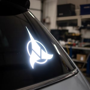 May include: A white, illuminated Starfleet emblem sticker on a car's rear window. The design includes an arrowhead, diamond, and circle. The sticker is placed on a dark, reflective surface.