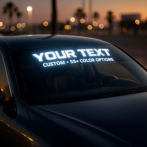 May include: A black car with a white illuminated windscreen banner displaying "YOUR TEXT" and "CUSTOM • 55+ COLOR OPTIONS". The background features a sunset with palm trees and blurred lights.