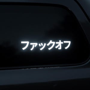 Puede incluir: Adhesivo de vinilo blanco con caracteres japoneses, que dice "ファックオフ", aplicado a una ventana de coche. El diseño es una declaración simple y audaz, adecuado para vehículos.