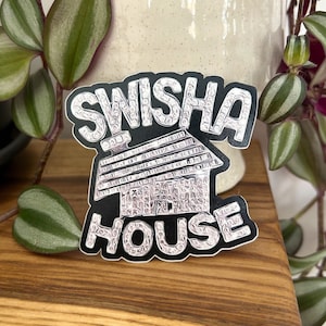 Könnte beinhalten: Ein schwarz-weißer Aufkleber mit den Worten "Swisha House" in einem rautenförmigen Muster. Der Aufkleber zeigt ein stilisiertes Hausdesign.