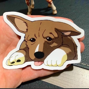 May include: A sticker depicting a cartoon corgi dog with brown and white fur, large ears, and a pink tongue. The dog is lying down with its paws forward.