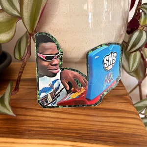 May include: A sticker featuring a cartoon image of a person wearing sunglasses and a white shirt, using a blue and red laptop with the text "SHS" on the screen.