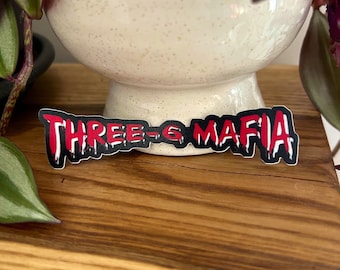 May include: A black and red sticker with the text "THREE-6 MAFIA" in a dripping font.