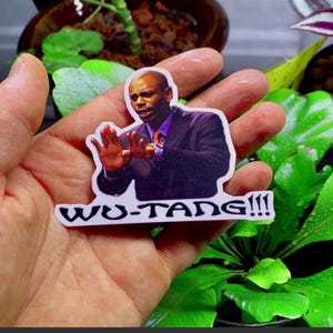 Puede incluir: Una pegatina blanca con el texto "WU-TANG!!!" y la imagen de un hombre con traje. La pegatina se sostiene en una mano, con plantas verdes en el fondo.