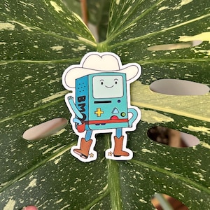 以下が含まれることがあります： カウボーイハットとブーツを履いた漫画のキャラクターのステッカー。キャラクターは、画面、赤いストラップ、文字「BMO」が付いた青い長方形です。ステッカーは白い縁取りで、緑の葉の上にあります。