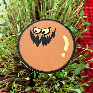 Op de afbeelding: Een ronde sticker met een cartoon monstergezicht. De sticker heeft een bruine achtergrond met een zwarte omtrek. Het monster heeft gele ogen en een grillige zwarte mond. Een geel uitroepteken staat aan de rechterkant.