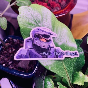 Può includere: Un adesivo con un personaggio della serie di videogiochi Halo che indossa un casco blu e viola. Il personaggio tiene una pistola e il testo "Wait. That's illegal." è stampato sotto il personaggio.