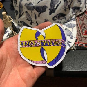 Op de afbeelding: Een gele Wu-Tang Clan sticker met een paarse en roze omtrek. Het Wu-Tang logo staat in het midden van de sticker. De sticker wordt in een hand gehouden.