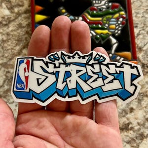 May include: A white and blue sticker with the word "STREET" in graffiti style, featuring a crown and the NBA logo. The sticker has a black outline and a blue shadow effect.