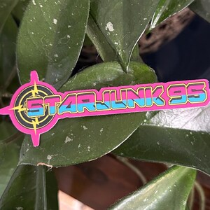 May include: A pink, black, and blue Star Junk 95 sticker. The sticker features the text "STAR JUNK 95" in a retro font, with a target design on the left side. The sticker is placed on a green leaf.