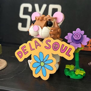 Puede incluir: Una figura de hámster Lego marrón con orejas rosas y mejillas blancas sostiene un letrero amarillo con las palabras "De La Soul" y una flor azul. A la derecha hay una flor morada con un tallo verde.