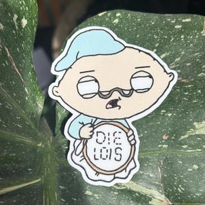 Op de afbeelding: Een sticker met een cartoonbaby met een blauwe nachtmuts en pyjama, met een bril en een verbaasde uitdrukking. De baby houdt een bord vast met de tekst "DIE LOIS" in een cirkelvormig ontwerp. De sticker heeft een witte rand.