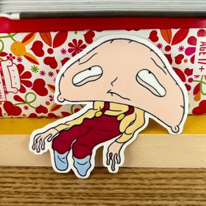 Op de afbeelding: Een sticker van een cartoonfiguur met een groot hoofd en een droevige uitdrukking. Het figuur draagt een rode overall en blauwe schoenen. De sticker zit op een boek met een bloemenpatroon.