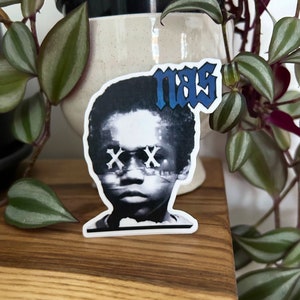 Op de afbeelding: Een zwart-witte sticker van een jong persoon met hun ogen bedekt met X'en. De sticker heeft het woord "mas" in blauwe letters boven het hoofd van de persoon.