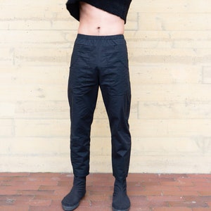 May include: A person is pulling a color-block sweater over their head. The sweater is black, tan, and black. They are wearing black pants and black shoes. The background is a light yellow wall.