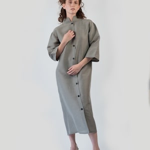 May include: A light gray midi dress with a mandarin collar and button-down front. The dress has three-quarter sleeves and is paired with white strappy heels. The fabric appears to be a textured linen or cotton blend.