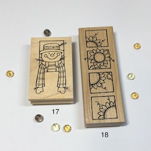 May include: Two rectangular wooden rubber stamps. Stamp 17 features a line art illustration of a scarecrow in a plaid shirt. Stamp 18 features a vertical design of four square panels, each containing a partial sunflower illustration. The number 17 is printed below the first stamp, and the number 18 is printed below the second.