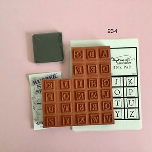 Könnte beinhalten: Ein Satz Alphabet-Stempel aus Gummi in einem braunen Holzrahmen. Die Stempel sind in einem Raster angeordnet und jeder Stempel zeigt einen Buchstaben des Alphabets in einer quadratischen Form. Die Stempel sind mit "Rubber Stamps Set" beschriftet.