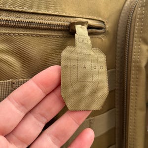 May include: Tan tactical backpack with a zipper and a target-shaped pull tab. The pull tab has the letters "A", "D", "C", and "G" printed on it. The backpack has multiple straps and a textured surface.